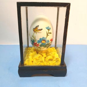 May include: A decorative egg with a butterfly and floral design, displayed in a glass and wood case. The egg rests on a bed of yellow fabric. The case has a black wooden base and frame.