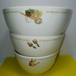 May include: A stack of three white ceramic bowls. Each bowl features a garden-themed illustration and the text "NATURE GIVES US THE VARIETY WE CRAVE." The illustrations include a wheelbarrow, gardening tools, and a watering can.