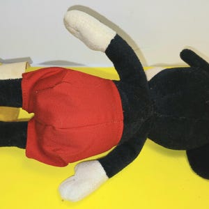 May include: A plush toy character featuring a black head and body, white hands, and bright yellow shoes. The character is wearing red shorts and has a small white fabric tag attached to the side of the left leg. The plush is positioned horizontally.
