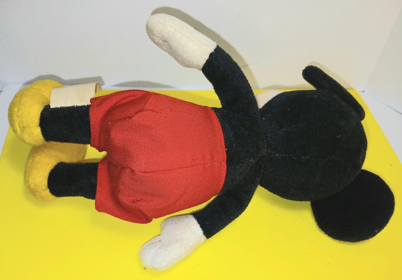 May include: A plush toy character featuring a black head and body, white hands, and bright yellow shoes. The character is wearing red shorts and has a small white fabric tag attached to the side of the left leg. The plush is positioned horizontally.