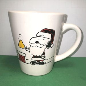 Peut inclure: Un mug en céramique blanche orné d'une illustration en noir et blanc du personnage Snoopy déguisé en Père Noël avec un bonnet rouge, une barbe blanche et un costume rouge, tenant une cloche jaune près d'une petite cheminée en briques rouges.