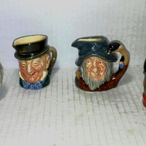 May include: Four miniature ceramic jugs, each sculpted to resemble a different man's face. The jugs feature detailed facial expressions, hats, and handles. The colour palette includes shades of brown, blue, and cream.