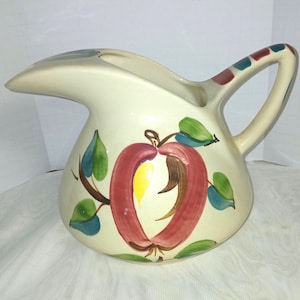 May include: Cream-colored ceramic pitcher with a large red apple design, green leaves, and brown stems. The handle has red and teal accents. The pitcher has a curved spout and a rounded body, perfect for serving beverages.