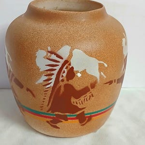 May include: A tan ceramic vase with a speckled texture featuring a dark brown silhouette of a person wearing a headdress and holding a white animal figure. The base is decorated with horizontal green, red, and yellow stripes around the circumference.