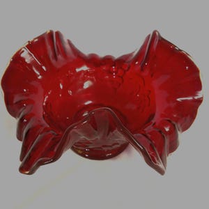 May include: A deep red glass bowl featuring a ruffled, crimped edge and a molded relief pattern of grapes and leaves on the exterior surface. The bowl has a wide, flared opening and a circular base, showcasing a translucent, vibrant ruby color.