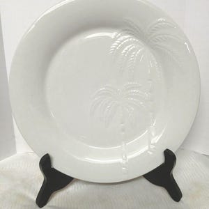 May include: A white ceramic plate with a raised palm tree design. The plate is round and sits on a black display stand. The palm tree design is visible on the right side of the plate.