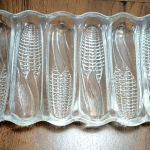 May include: Clear glass serving dish with six corn-shaped compartments. The dish has a rectangular shape with handles on each end. The words "Maize" are visible on the side. A vintage kitchenware item.