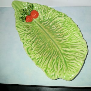 May include: A ceramic serving dish shaped like a large, textured green lettuce leaf. The dish features a prominent central vein and a decorative cluster of two small, bright orange radish-like vegetables with green leaves at the top end.