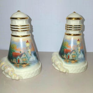 Può includere: Un paio di saliere e pepiere a forma di faro. I shaker in ceramica sono bianchi con finiture dorate e presentano una scena costiera dipinta con un faro, una casa e onde oceaniche. La base ha un design a corda.