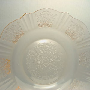 May include: A pale pink translucent glass plate featuring a scalloped edge and a central circular medallion with intricate embossed floral and scroll patterns. The rim is decorated with repeating embossed motifs that match the center design.