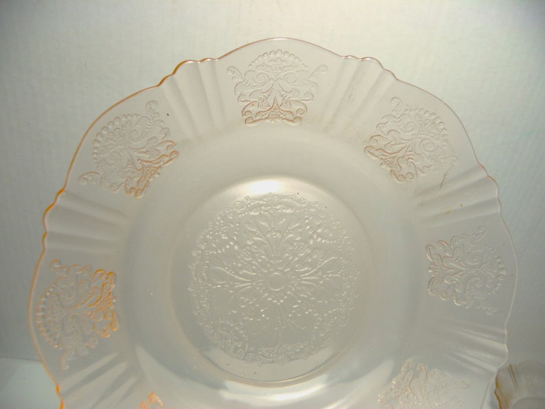 May include: A pale pink translucent glass plate featuring a scalloped edge and a central circular medallion with intricate embossed floral and scroll patterns. The rim is decorated with repeating embossed motifs that match the center design.