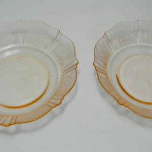 May include: Two matching shallow bowls made of translucent pale orange glass. Each bowl features a scalloped edge and a delicate embossed floral pattern around the rim and center, with a smooth, circular base and a slightly flared, curved side profile.