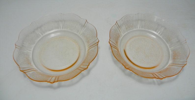 May include: Two matching shallow bowls made of translucent pale orange glass. Each bowl features a scalloped edge and a delicate embossed floral pattern around the rim and center, with a smooth, circular base and a slightly flared, curved side profile.