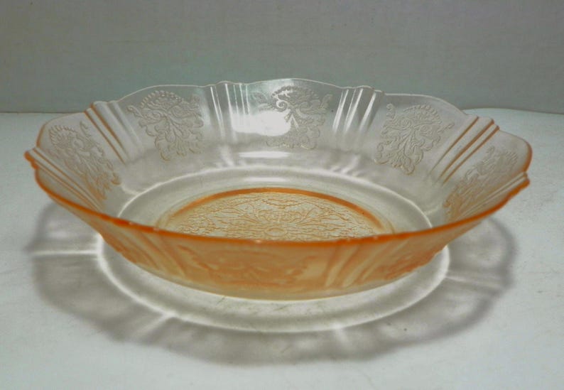 May include: A shallow, round, peach-colored glass bowl featuring a scalloped rim and a textured, embossed floral pattern around the interior sides and a detailed, intricate geometric design centered on the bottom of the basin.