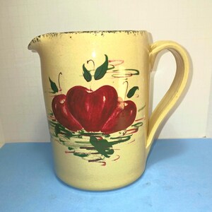 May include: A pale yellow ceramic pitcher featuring a hand-painted design of three red apples with green leaves. The pitcher has a curved handle, a pouring spout, and a speckled texture along the top rim, with visible brushstrokes throughout.