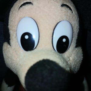 May include: A plush toy of the character Mickey Mouse featuring a cream-colored face, a large black nose, and prominent black ears. The character has large, white, oval-shaped eyes with black pupils and a soft, textured fabric construction.