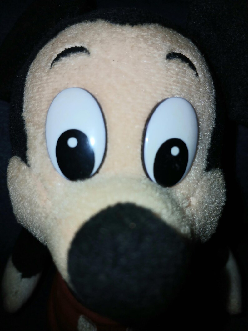 May include: A plush toy of the character Mickey Mouse featuring a cream-colored face, a large black nose, and prominent black ears. The character has large, white, oval-shaped eyes with black pupils and a soft, textured fabric construction.