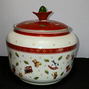 May include: A white ceramic jar with a red lid and accents, decorated with Christmas-themed illustrations. The jar features images of teddy bears, trains, rocking horses, and holly leaves. The lid is topped with a green finial.