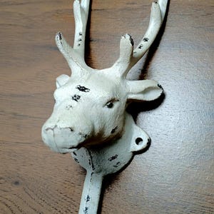 May include: A white cast iron deer head wall hook with a distressed finish. The deer head features detailed antlers and a hook for hanging items. The hook is mounted on a wooden surface.