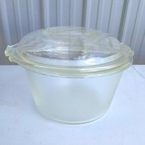 May include: A clear glass bowl with a matching lid. The bowl has a frosted appearance, while the lid is transparent. The lid features a ridged design on top and small handles on either side. The bowl is likely for food storage or serving.