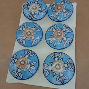 May include: Six round ceramic cabinet knobs. Each knob has a light blue background with a white floral design and a gold-coloured centre. The knobs are arranged on a white surface.