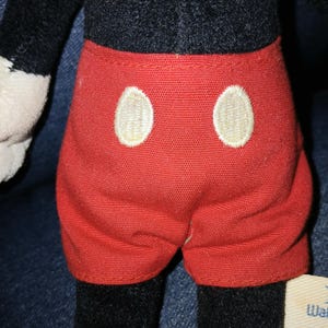 May include: A plush toy featuring a black torso and bright red shorts with two large, oval-shaped white buttons embroidered on the front. The toy has black limbs and a small white fabric tag attached to the side with visible text.