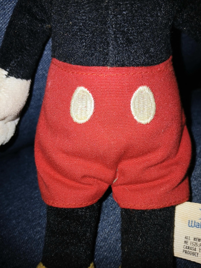May include: A plush toy featuring a black torso and bright red shorts with two large, oval-shaped white buttons embroidered on the front. The toy has black limbs and a small white fabric tag attached to the side with visible text.