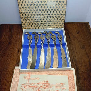 May include: A set of six silver butter knives with seahorse-shaped handles, presented in a decorative box. The box has a blue interior and a patterned lid. The set includes a card with text that reads "This exquisite 6-piece set of silver from Florence Italy."
