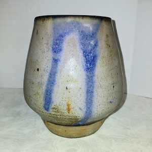 May include: A handmade ceramic vase with a unique, organic shape. The vase features a speckled, off-white base with vertical streaks of blue glaze. The rim is dark brown, and the base is a lighter tan color.