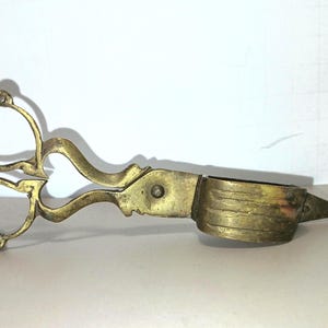 May include: A brass wick trimmer with ornate, curved handles and a cylindrical cup-shaped head. The metal has a weathered, aged patina with visible texture and a pointed tip extending from the end of the circular cutting mechanism.