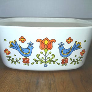 May include: White rectangular casserole dish with handles, decorated with blue birds, orange flowers, and green foliage. The vintage dish is likely used for baking and serving food.