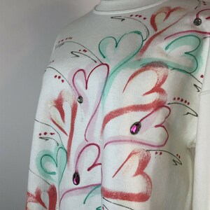 May include: White sweatshirt with a colorful abstract design featuring hearts, leaves, and swirls. The design is painted in shades of pink, red, green, and white. The sweatshirt is decorated with small pink rhinestones.