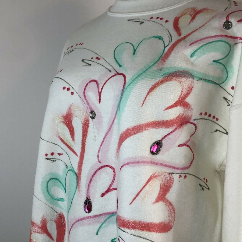 May include: White sweatshirt with a colorful abstract design featuring hearts, leaves, and swirls. The design is painted in shades of pink, red, green, and white. The sweatshirt is decorated with small pink rhinestones.