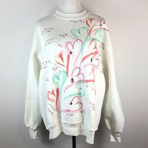May include: White sweatshirt with a colorful abstract floral design featuring pink, red, green, and blue accents. The design includes several small rhinestones.