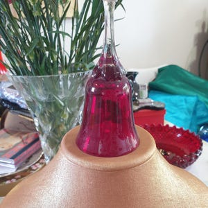 Puede incluir: Una botella de vidrio rojo rubí con cuello largo y base acampanada. Descansa sobre un expositor dorado. Claveles rosas en un jarrón transparente al fondo. El tema general es cristalería decorativa.