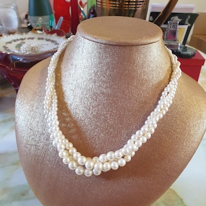 Puede incluir: Un collar de perlas de varias hebras expuesto sobre un busto dorado. El collar presenta tres hebras de perlas blancas y redondas. El busto es de color dorado sólido, y el fondo está ligeramente borroso, con indicios de otros objetos.