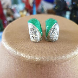 May include: A pair of silver-coloured clip-on earrings. Each earring has a curved, emerald green enamel top and a section encrusted with clear, round rhinestones. The earrings are displayed on a tan surface.