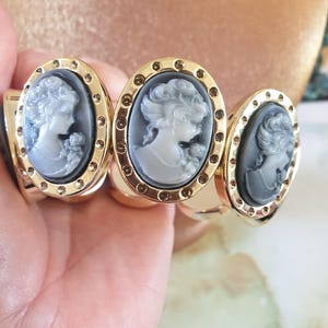 Puede incluir: Un brazalete de oro con tres dijes ovalados. Cada dije presenta un camafeo de una mujer con la cara blanca y el pelo negro. Los dijes están rodeados por una fila de pequeños puntos dorados.