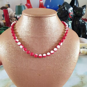 May include: A red and white beaded necklace on a gold display stand. The beads are alternating red and white.