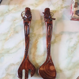 Könnte beinhalten: Ein Set aus zwei hölzernen Salatbesteck mit geschnitzten Gesichtern und eingelegten Türkis-Steinen. Die Bestecke sind braun und haben eine natürliche Holzoptik.