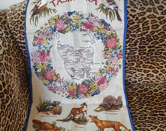 Puede incluir: Un paño de cocina de lino con un mapa de Tasmania rodeado por una corona floral. La palabra "TASMANIA" está en la parte superior. Ilustraciones de animales nativos, incluyendo un ornitorrinco, un canguro y un tigre de Tasmania, en la parte inferior. Borde azul.