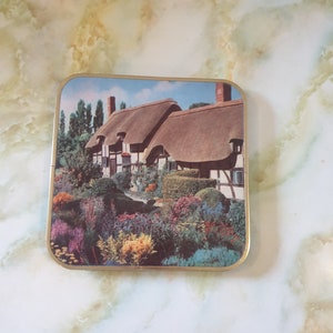 May include: A vintage tin featuring a colourful image of a thatched-roof cottage surrounded by a lush garden with flowers in shades of pink, purple, yellow, and blue.