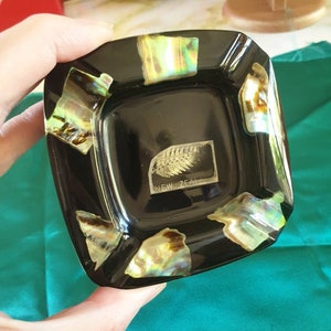 May include: Black ashtray with a square shape and a raised center. The ashtray is decorated with iridescent mother-of-pearl accents and features a fern leaf design with the text "NEW ZEALAND".
