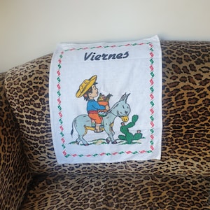 May include: White kitchen towel with a red, green and white border. The towel features a cartoon illustration of a person riding a donkey with a cactus in the background. The text "Viernes" is printed in black at the top of the towel.