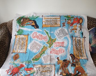 Op de afbeelding: Een vintage witte en blauwe theedoek met een kaart van Nieuw-Zeeland en de woorden "Cooking Kiwi Style". De theedoek bevat illustraties van eten, mensen en tekst over kookmethoden.