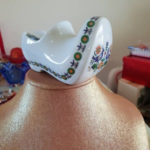 Könnte beinhalten: Weißer Keramik-Schuh als Dekorationsobjekt mit einem grünen, orangefarbenen und blauen Blumendesign. Der Schuh steht auf einem goldfarbenen Präsentationsständer.