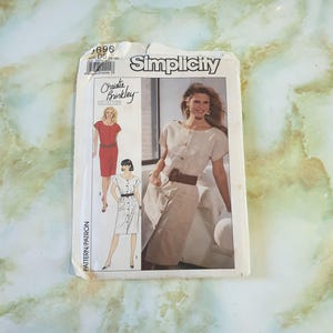 Pode incluir: Padrão de costura Simplicity com um design da Coleção Christie Brinkley. O pacote mostra ilustrações de vestidos, incluindo um vestido abotoado. O pacote é branco com o logotipo Simplicity.