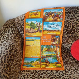 May include: A vintage orange tea towel with a colorful illustration of scenes from the Australian outback. The scenes include a flock of sheep, a horse and rider, a wheat field, and a river. The text "Greetings From Bowral" and "The Australian Outback" are printed on the towel.