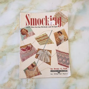 May include: A vintage book titled "Smocking: New Time-Saving Methods and Designs" by Irene A. Noll. The cover features illustrations of smocking designs and a woman sewing.