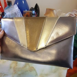 Bolso de mano o bandolera de los años 80. Pvc plateado, dorado y bronce con diseño de inspiración art déco. Correa larga desmontable.